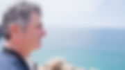 Homem adulto de cabelos grisalhos à esquerda da imagem em grande plano, em background, à distância vista de mar. 