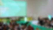 Pessoas assistem a uma palestra. No palco 4 pessoas sentadas e atrás deles um ecrã gigante com o nome do evento.