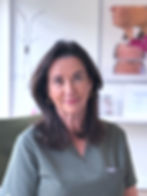 Smiling woman in green FIGS scrubs indoors, with dark hair and hoop earrings, standing in a bright room.
