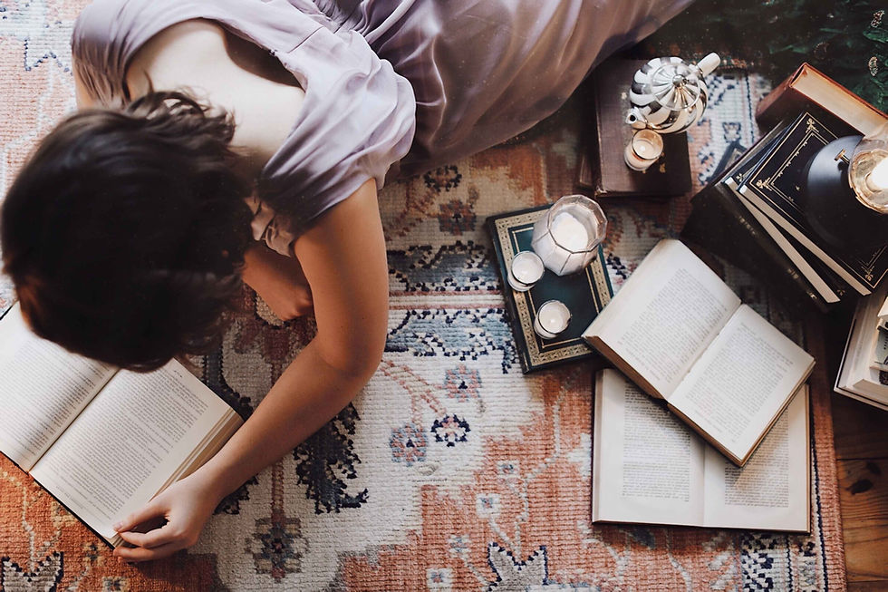 Woman in a lavender dress reads on a patterned rug, surrounded by open books, a candle, and a teapot, in a calm, creative and cozy scene