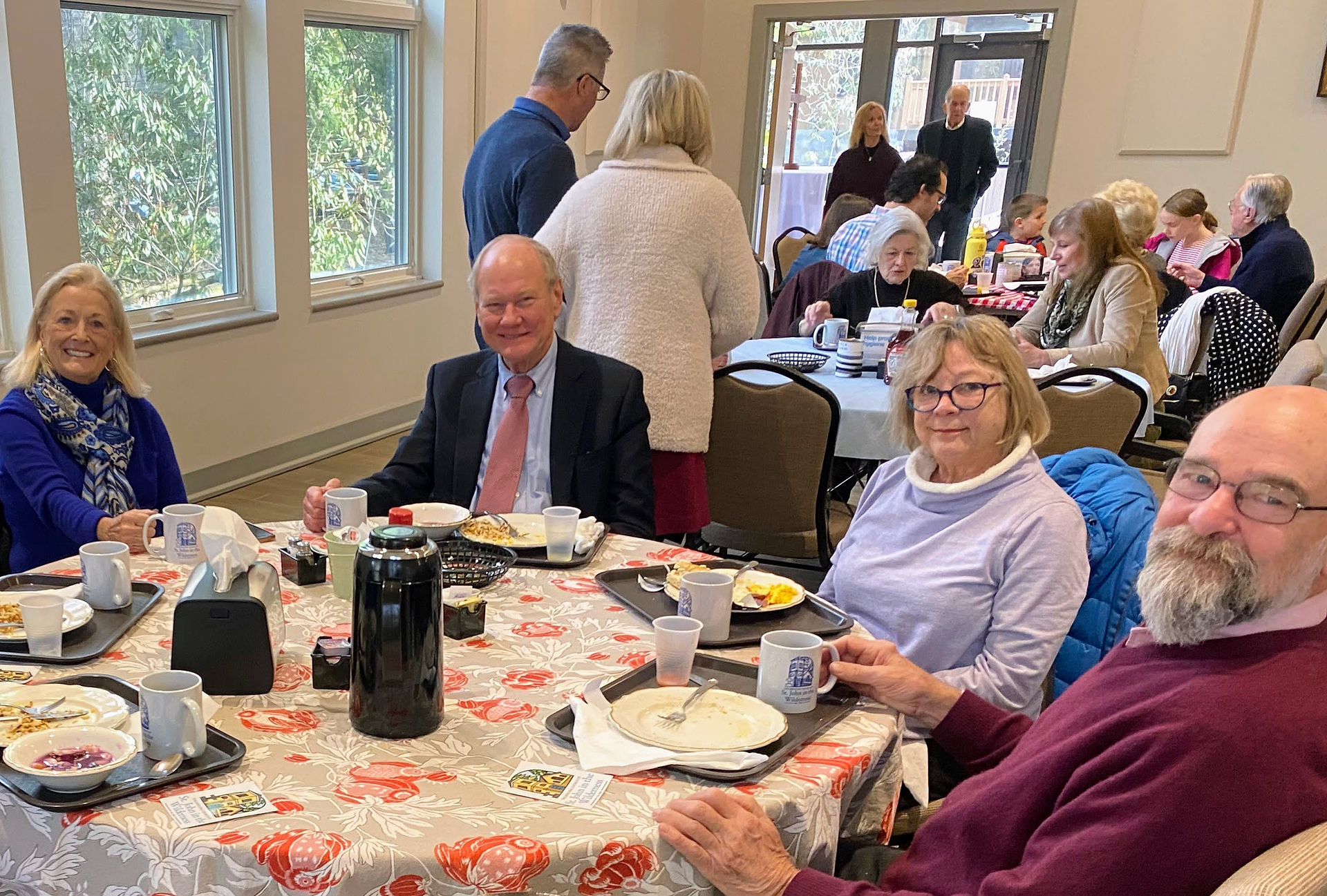 Four people sit in the foreground around a breakfast table, smiling with plates of food in front of them. In the background, more people gather around tables and converse as they enjoy their meals.