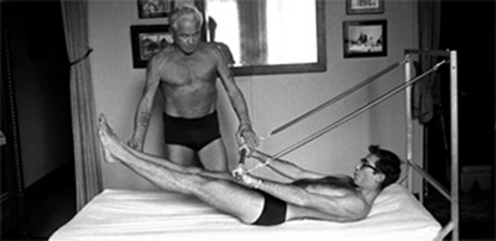 Historisches Schwarz-Weiß-Foto: Joseph Pilates korrigiert einen Schüler, der auf einem Trapeze Table mit Federn trainiert
