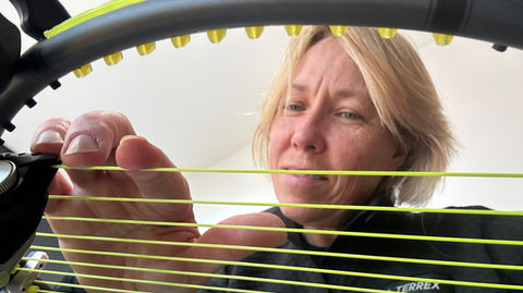 A person is looking over a tennis racquet and pressing her finger on the main strings which is mounted on a  stringing machine