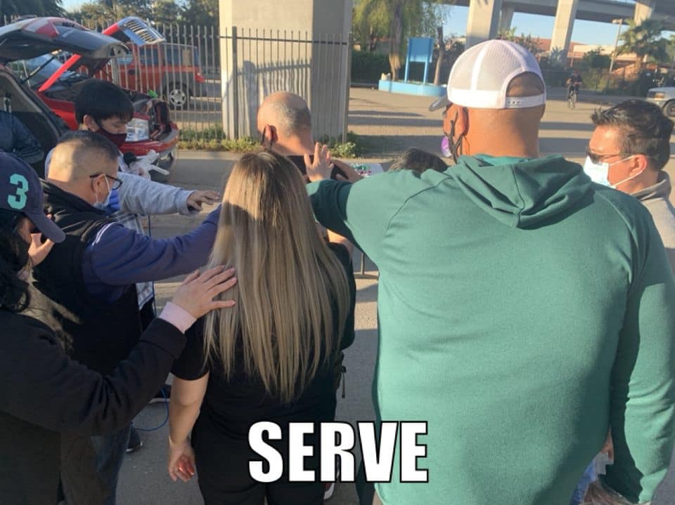 A diverse group of volunteers standing together in a circle with arms around each other, praying outdoors