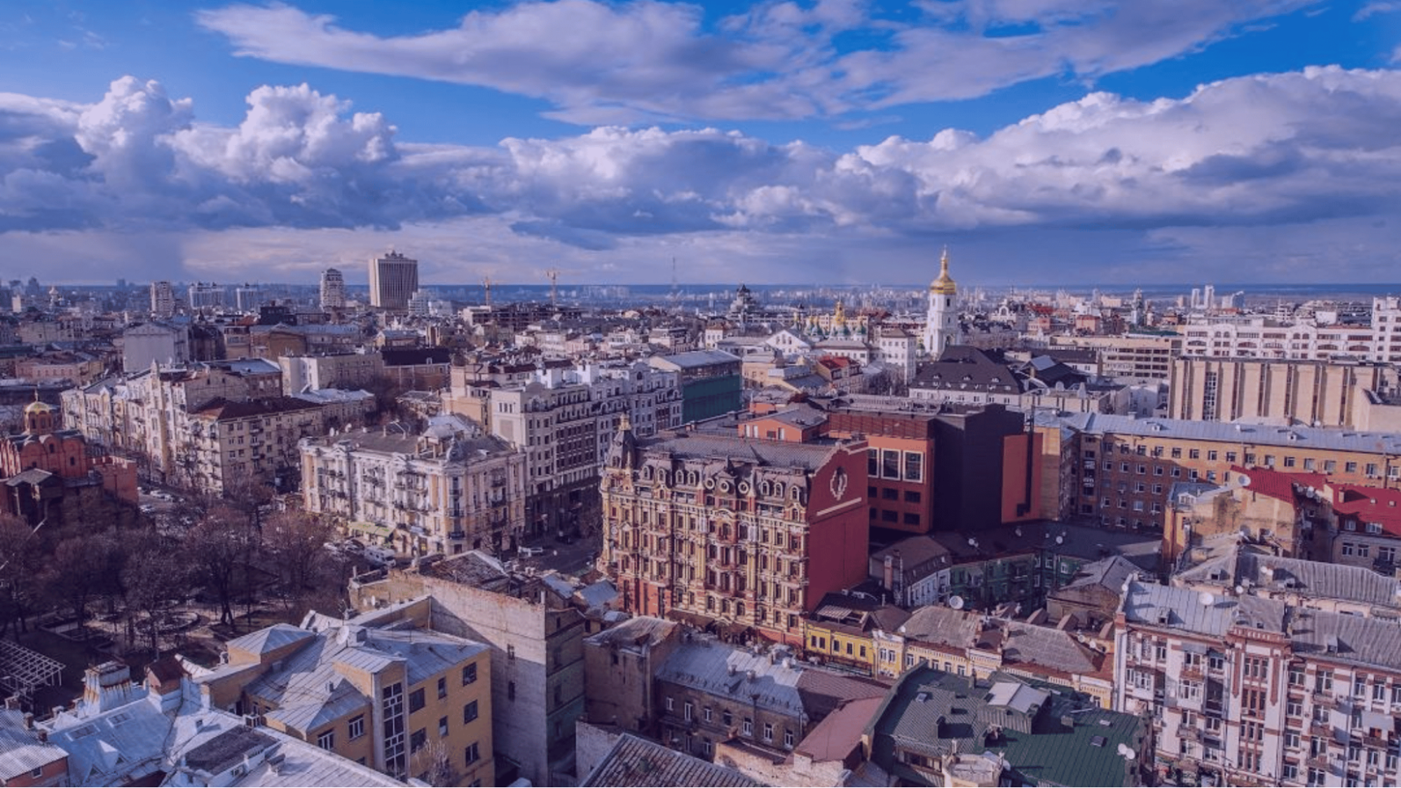 Aerial view of a city with diverse architecture under a blue, cloudy sky. Prominent buildings include red and white structures and a golden dome.