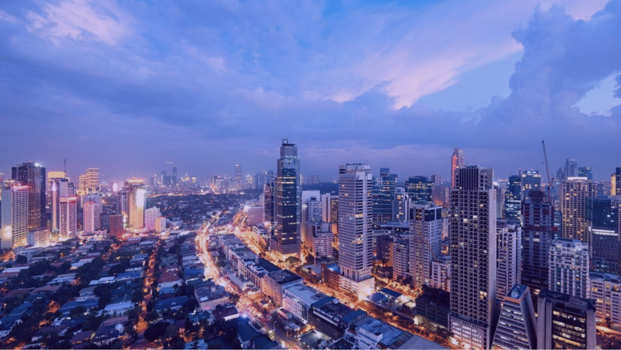 City skyline at dusk, illuminated buildings with vibrant lights, under a partly cloudy blue and purple sky. Streets are busy with traffic.