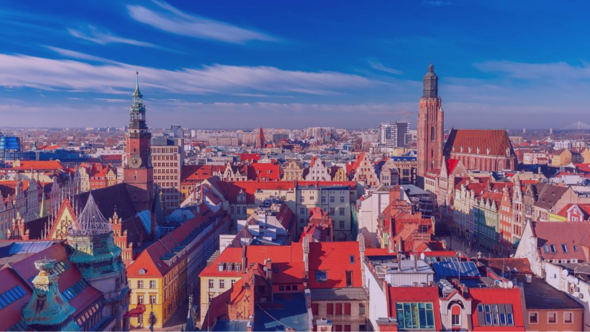 Cityscape with colorful, red-roofed buildings and tall church towers under a bright blue sky. Vibrant urban atmosphere, no text visible.