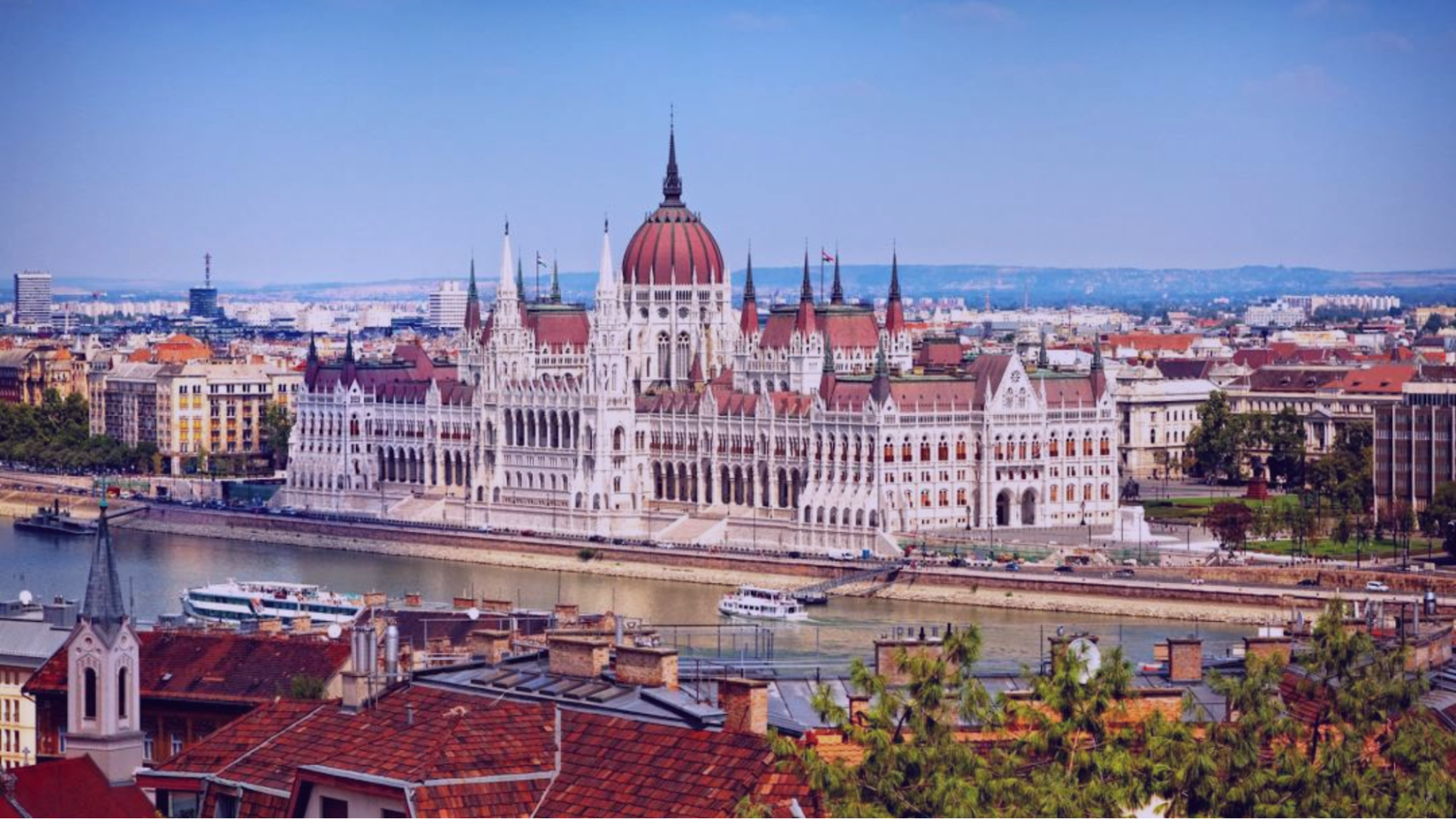An ornate Gothic building with red roofs by a river under a clear blue sky, surrounded by a cityscape and lush greenery. Boats visible.