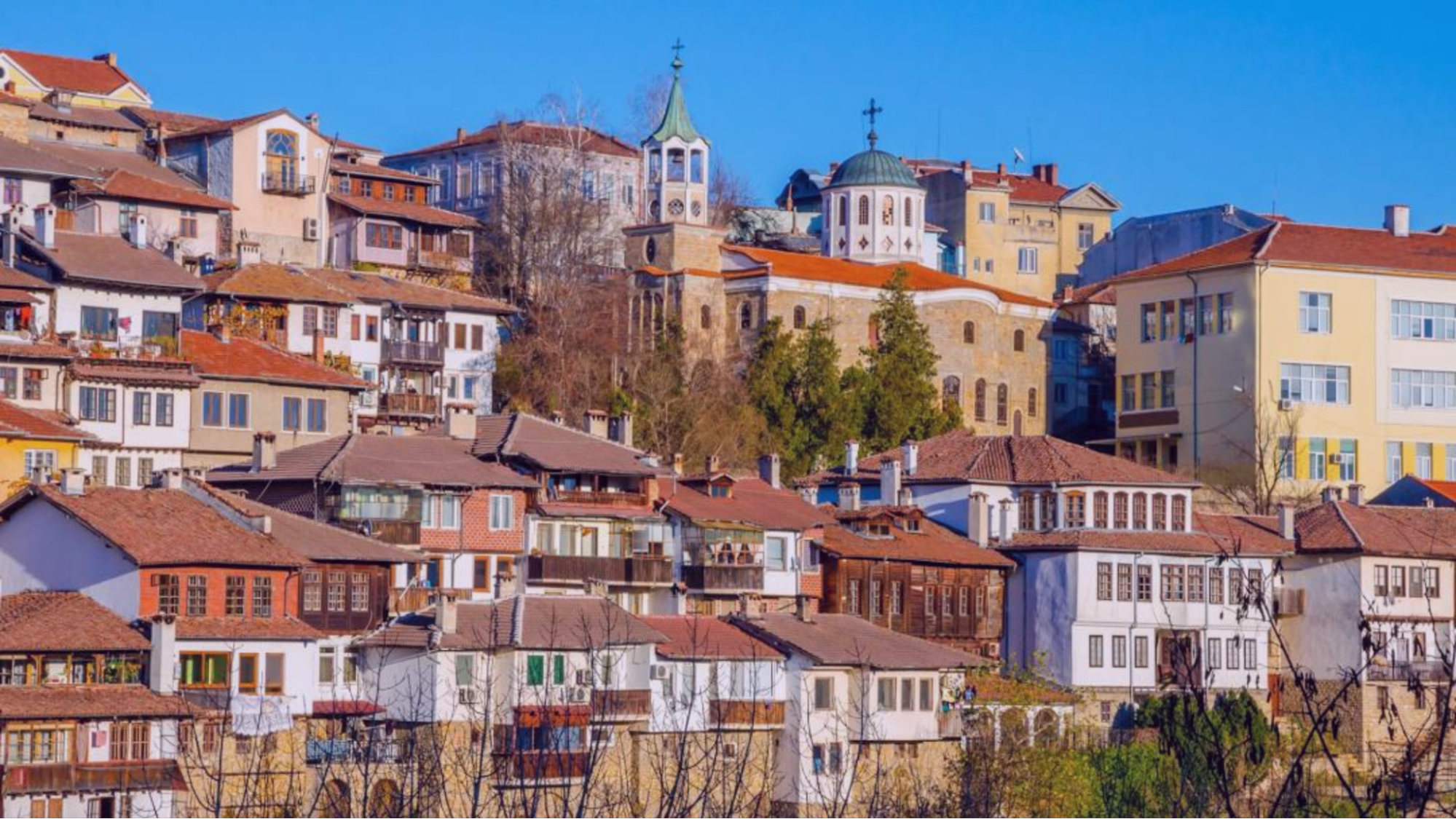 Colorful hillside town with red-roofed houses and a church with domes and cross. Clear blue sky above. Vibrant and serene atmosphere.