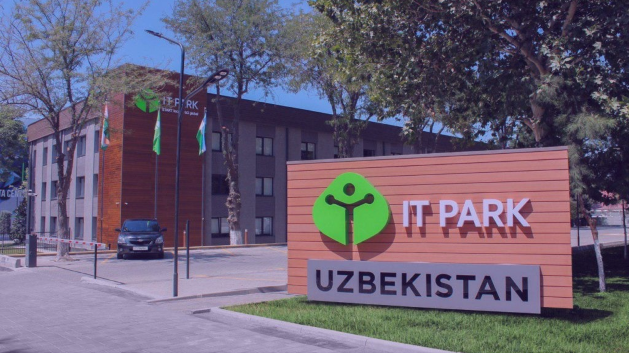 Sign reads "IT Park Uzbekistan" with green logo. Modern building, car, and flags in background. Trees and clear blue sky set a serene scene.