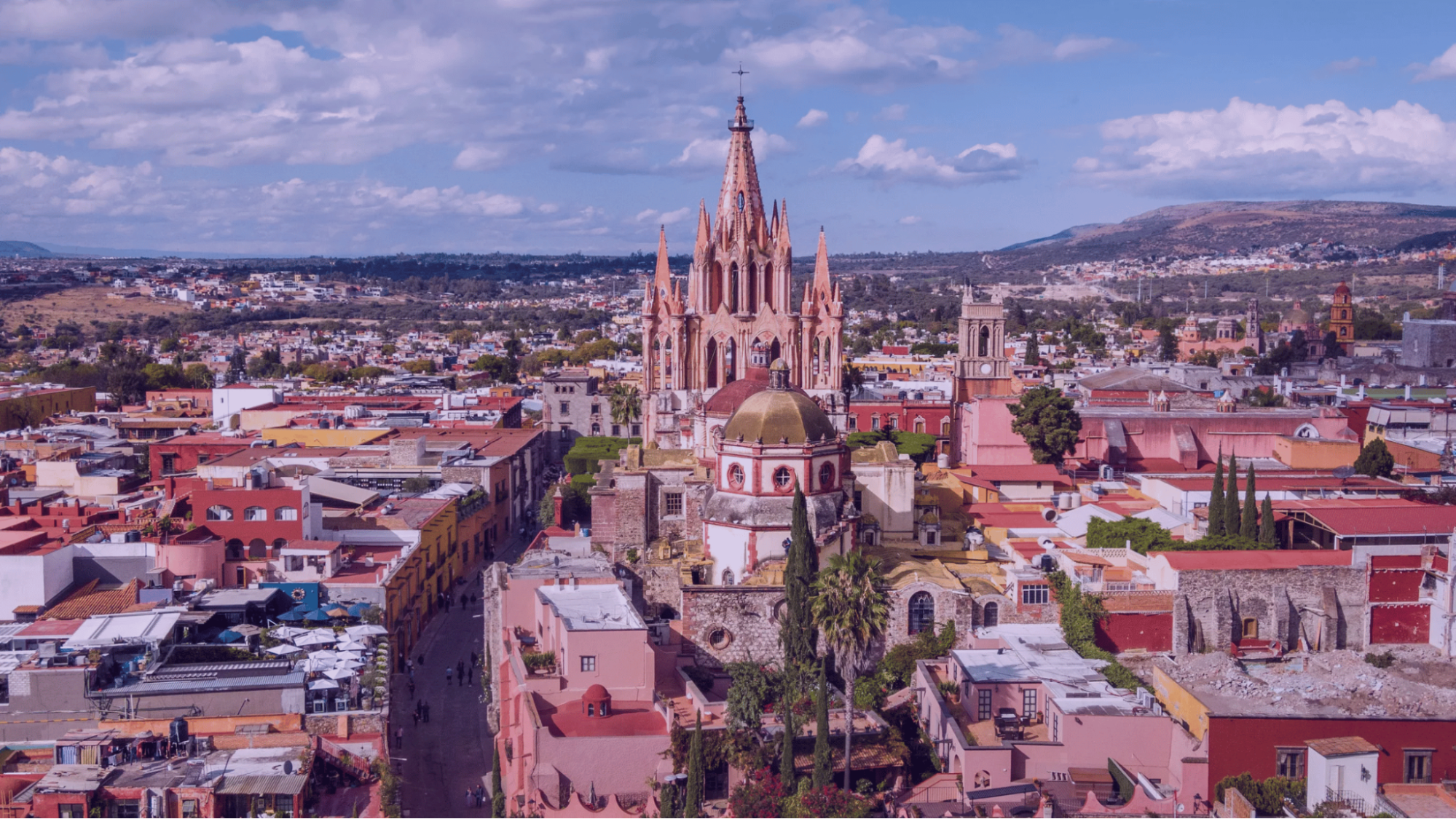 Aerial view of a colorful colonial city with a pink cathedral spire, domed rooftops, hills, and cloudy blue sky.