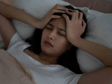 Woman lying in bed with eyes closed, clutching her head in pain, surrounded by white pillows and beige bedding.