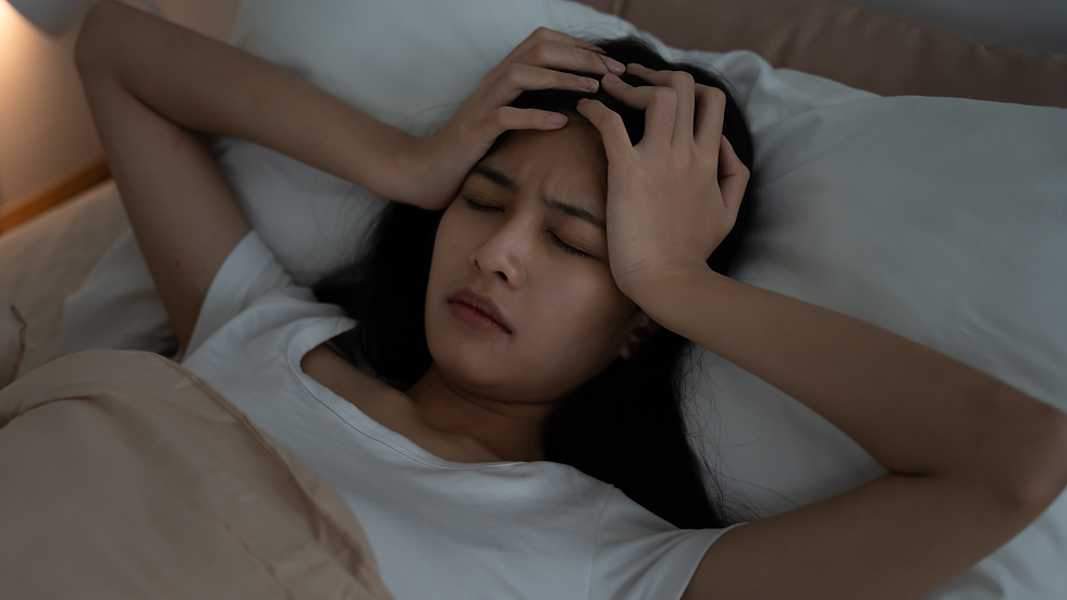 Woman lying in bed with eyes closed, clutching her head in pain, surrounded by white pillows and beige bedding.