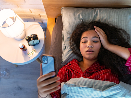 Young woman in red striped pajamas леж? no, in bed holding her head and looking at a phone beside a lit lamp, pill bottle, and alarm clock