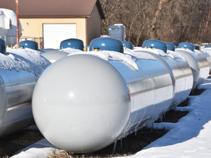 White propane tanks covered in snow, aligned outdoors with a brown shed and bare trees in the background. Bright winter sunlight.