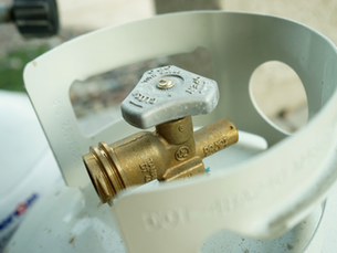Close-up of a propane tank valve with a brass body and silver handle. The background is slightly blurred with some text visible.