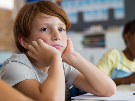 Thoughtful red-haired boy rests his face in his hands in a classroom, with a blurred classmate drawing behind him.