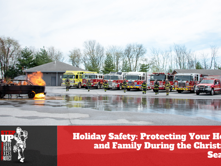 Firefighters in full turnout gear walk across a training ground toward a controlled vehicle fire. Several fire engines and emergency vehicles are lined up in the background, and flames rise from the car while water reflects on the pavement. A red banner at the bottom reads, “Holiday Safety: Protecting Your Home and Family During the Christmas Season,” alongside a “Step Up” firefighter recruitment graphic.