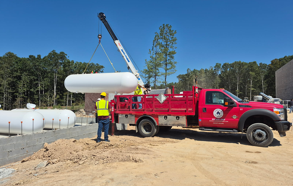 Workers in hard hats guide a large propane tank lifting by a crane from a red truck. Clear blue sky, forest, and construction site backdrop.