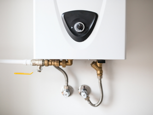 White tankless water heater with a black dial, brass fittings, and metal pipes against a plain wall. The setting is clean and minimal.