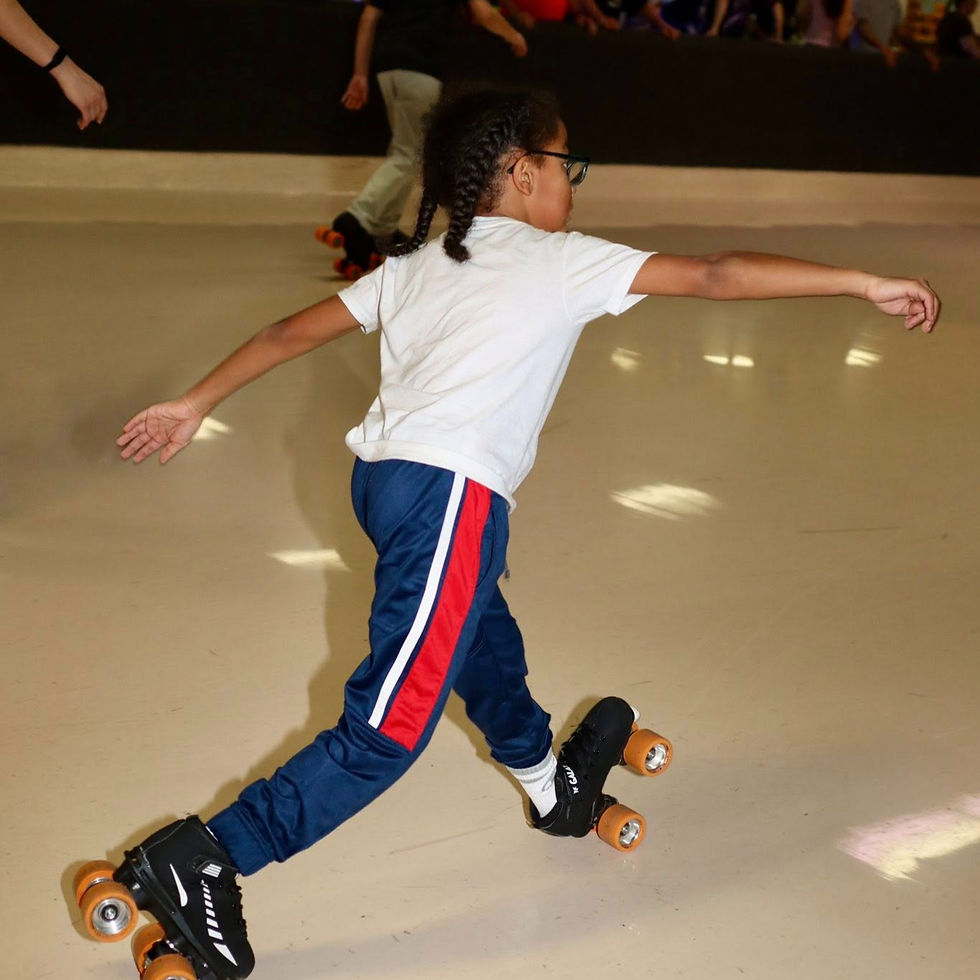 A child in braids and glasses roller skates indoors, arms outstretched. Wears a white shirt and blue pants with red stripes.