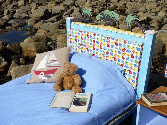 Bed on the beach with teddy bear and book; blue sheets and pillow.