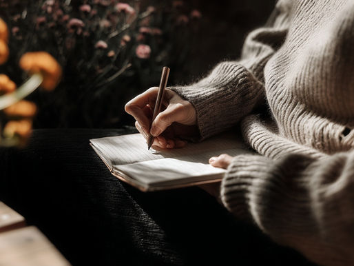 A person in a cozy sweater writes in a notebook, surrounded by orange flowers. Warm, soft lighting creates a calm, reflective mood.