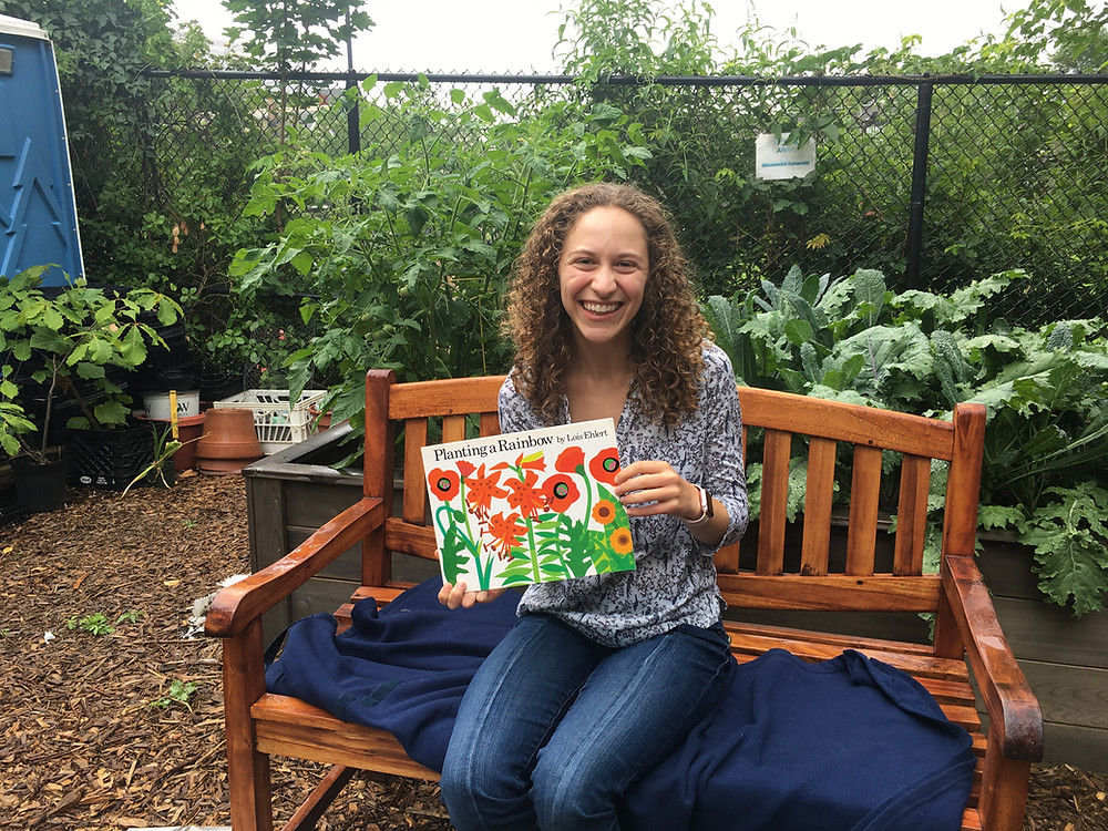Becca sitting on a bench in the raised bed section of the garden, holding the book, "Planting a Rainbow."