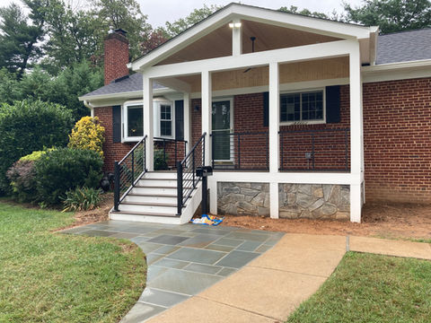 This is the street view of a cozy brick home with a freestanding porch framed in white with a black fence around the perimeter. There is hardscape path leading to 6 stairs that lead up to the porch.