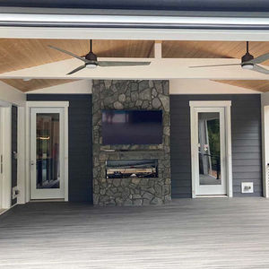 This is a back deck from inside the deck. There is a floor to ceiling fireplace made of large stones in the middle framed with two doors to inside the house. There is a rectangle fireplace embedded with a stone mantle above it and a mounted TV. Gray tones floors with lighter wood planks on the ceiling.