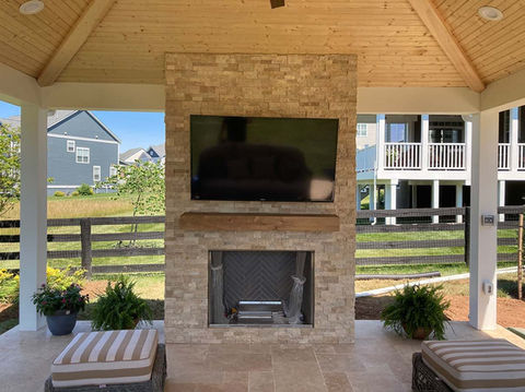 A-frame style indoor-outdoor porch. Customized to the client's preferences, a stone fireplace is in the center with a wood plank mantel and large square fireplace embedded. The ceiling is al light wood and the sides of the structure are open to allow for shade but still incorporate the Northern Virginia Landscape.