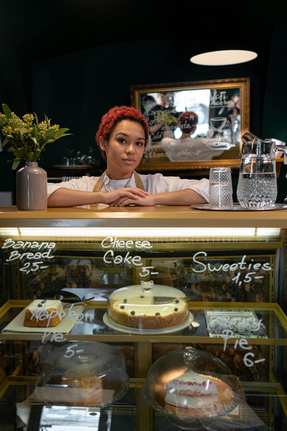 Woman standing behind a display case with a variety of baked goods with pricing on the display window