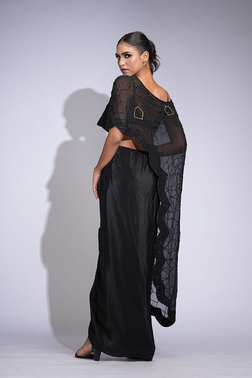 Back view of model showing flowing organza cape and asymmetrical pleats on black drape skirt in motion.