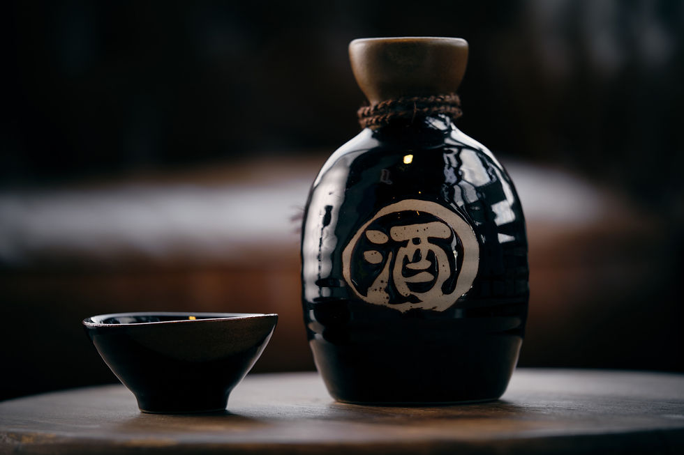 Dark ceramic sake bottle with Japanese characters, next to a matching cup on a wooden table. Dimly lit and cozy setting.