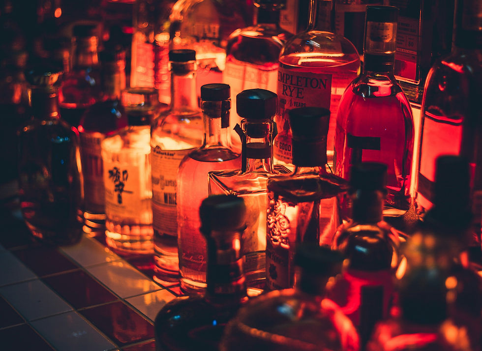 Rows of liquor bottles on a dimly lit bar emit warm, glowing amber tones. The background features blurry red and orange hues.