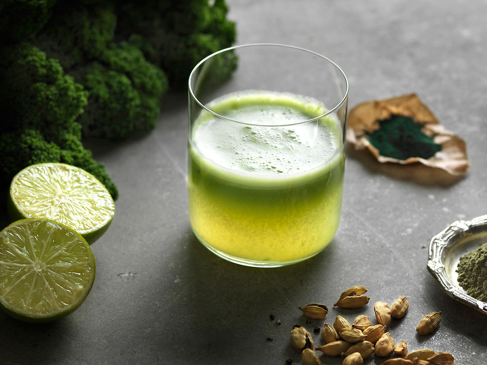 A glass of green juice on a gray surface surrounded by lime halves, cardamom pods, and green powders. Broccoli in the background.