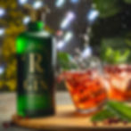 Green Revivallist gin bottle beside an orange cocktail with mint, ice, and pomegranate splashes on a wooden board.