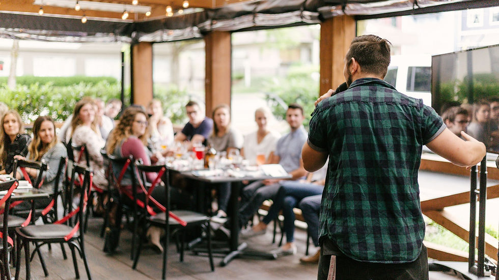 Man standing and speaking to a large group seated at long tables on a restaurant patio