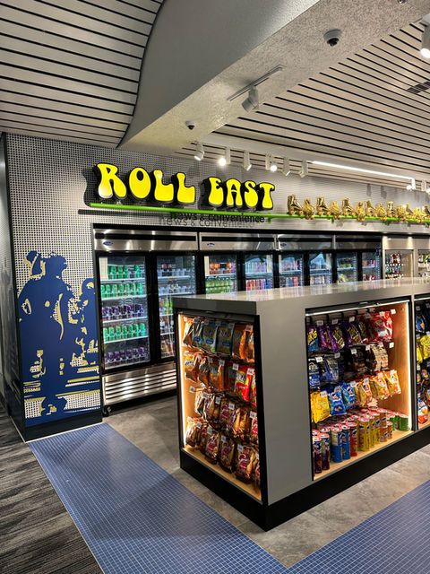 An airport convenience store with fridges stocked with cold drinks and a chip stand in the center.