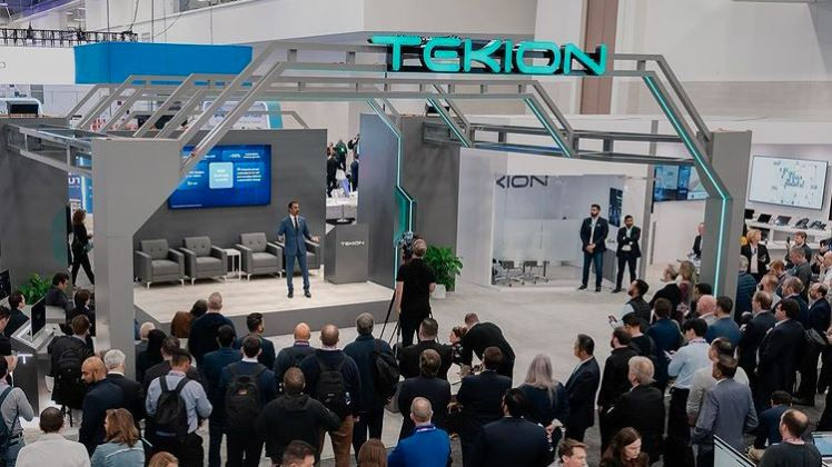 A man stands on a stage of a large trade booth and gives a presentation to a crowd of people.