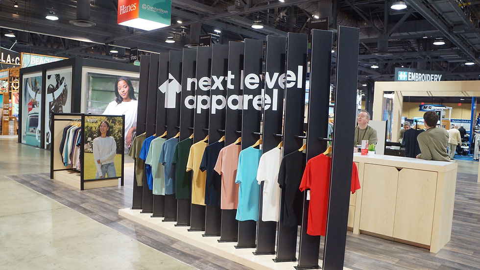 Display of colorful t-shirts on racks with "next level apparel" branding. Booth setting with people around, vibrant and inviting atmosphere.