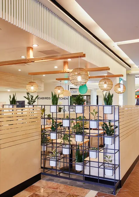 Interior detail showing slatted wood dividers, woven pendant lamps, and a metal plant wall with potted greenery inside Seabirds Kitchen.