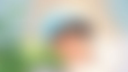 Child sleeping peacefully on a pillow, wearing a light blue hat with patterns and a green shirt. Soft pastel room with leafy plant nearby.