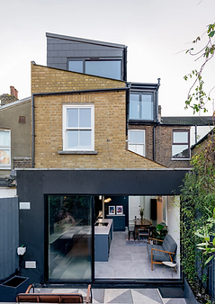 The image shows a modern three-story house with a brick façade and black cladding, featuring large glass doors that open into a sleek kitchen and living space. The upper floors have a contemporary, angular design, blending traditional and modern architecture.