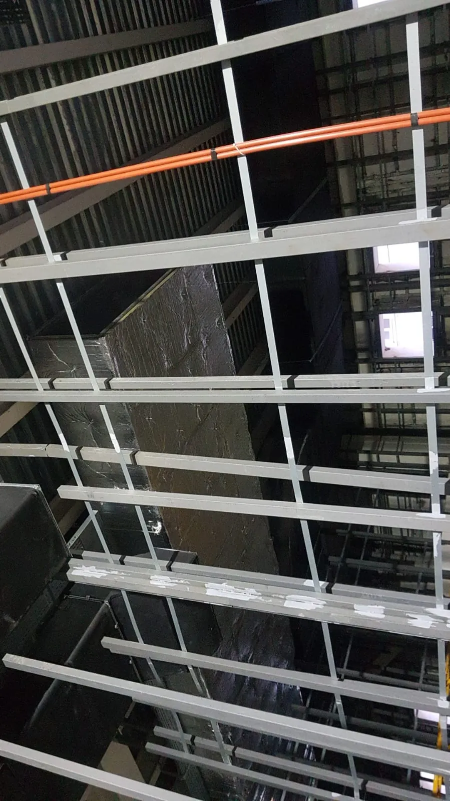 Asma tavan karkası üzerinde uzanan folyo kaplı dikdörtgen kanal, çelik taşıyıcılar ve turuncu borular