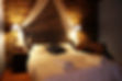 Large double bed with white bedcover, and folded white towels. A bedside table and bedside lamp to the side of the bed, with a chair next to it. The wall behind the bed is made from natural stone.