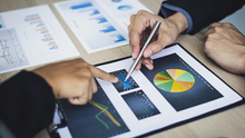 Hands in a business meeting point at charts and graphs on printed reports and a tablet, with a pen over a pie chart.