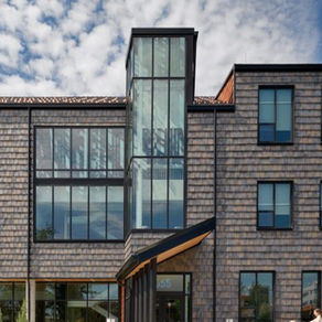 Modern gray-shingled building, Ronald McDonald House in Dayton, Ohio, with tall glass stair tower under a cloudy blue sky; a small person stands near the entrance.