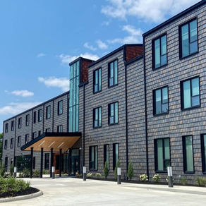 RMHC Dayton, a modern shingled building with tall windows and a glass entry canopy under a blue sky, surrounded by landscaped greenery.