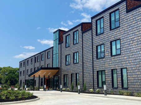 RMHC Dayton, a modern shingled building with tall windows and a glass entry canopy under a blue sky, surrounded by landscaped greenery.